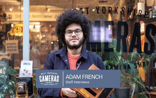A man with an afro and glasses standing in front of a camera shop.