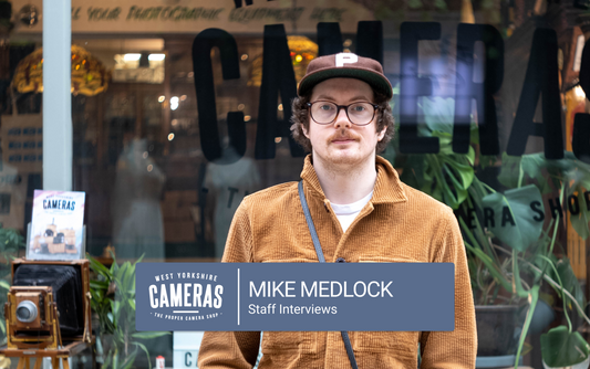 Man with a cap, moustache and glasses standing in front of a camera shop.